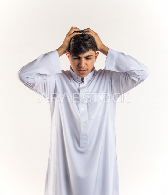 بورتريه شاب مراهق خليجي سعودي مريض يضع يده على رأسه بإيماءات وجه ويدين مختلفة تدل على الألم و الوجع ، خلفية بيضاء