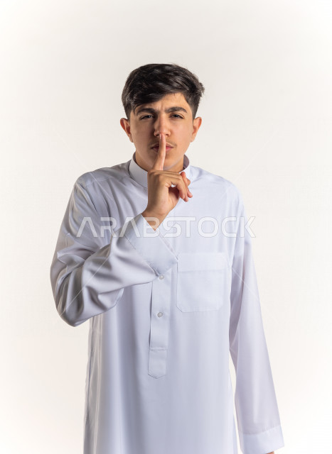Portrait of a young Saudi Arabian Gulf teenager pointing his finger at his mouth for silence and silence with different facial and hands gestures, white background