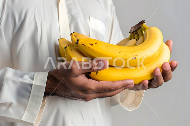 انتقاء أجود أنواع الفواكه الطازجة، بورتريه مقرب لأيدي شاب عربي خليجي سعودي يرتدي الثوب التقليدي يحمل بيده حبات الموز، المحاصيل الزراعية السعودية ذات الجودة العالية، وجبة طعام صحية خفيفة، تلبية الحاجات الضرورية للجسم، خلفية بيضاء