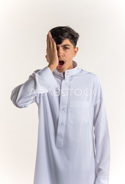 بورتريه شاب مراهق خليجي سعودي مريض بإيماءات وجه ويدين مختلفة تدل على الألم و الوجع في الأسنان ، يرتدي ثوب أبيض , خلفية بيضاء