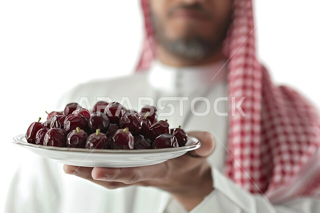 وجبة صحية ذات قيمة غذائية عالية, منتجات ومحاصيل زراعية محلية، بورتريه مقرب لرجل عربي خليجي سعودي يرتدي الشماغ و الثوب التقليدي يحمل بيده طبق من حبات التمر الطازجة, خلفيه بيضاء