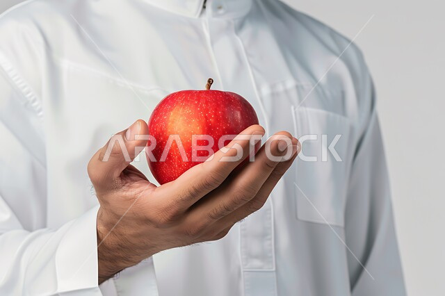 وجبة صحية ذات قيمة غذائية عالية, منتجات ومحاصيل زراعية محلية، بورتريه مقرب لرجل عربي خليجي سعودي يرتدي الثوب التقليدي يحمل بيده تفاحة حمراء اللون, المحافظة على اللياقة البدنية، خلفيه بيضاء