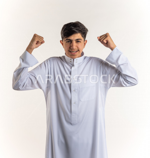 بورتريه شاب مراهق خليجي سعودي يقوم بحركة العضلات ذات الرأسين بإيماءات وجه ويدين مختلفة , خلفية بيضاء , التمتع بصحة بدنية جسدية جيدة