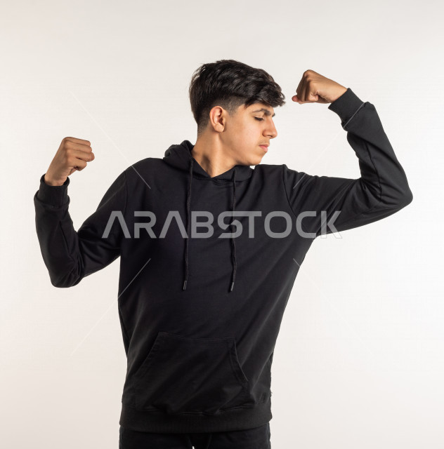 بورتريه شاب مراهق خليجي سعودي يقوم بحركة العضلات ذات الرأسين بإيماءات وجه ويدين مختلفة , خلفية بيضاء , التمتع بصحة بدنية جسدية جيدة