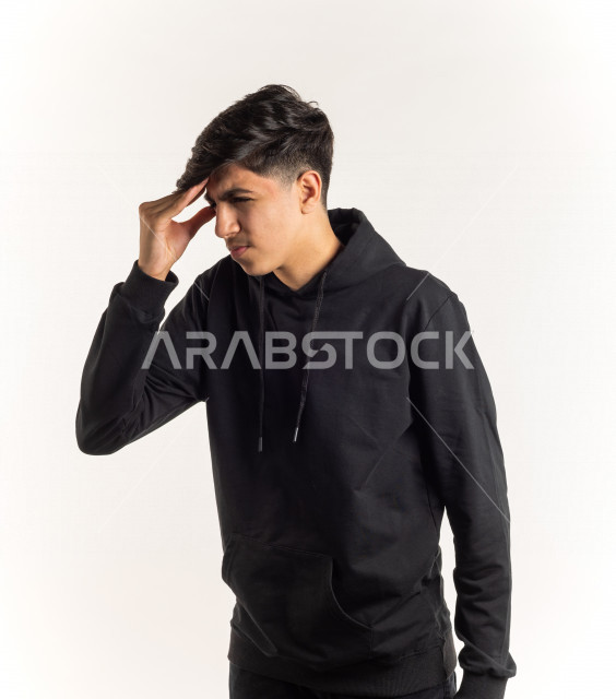 بورتريه شاب مراهق خليجي سعودي مريض يضع يده على رأسه بإيماءات وجه ويدين مختلفة تدل على الألم و الوجع ، خلفية بيضاء
