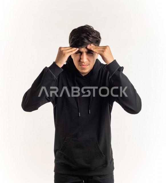 بورتريه شاب مراهق خليجي سعودي مريض يضع يده على رأسه بإيماءات وجه ويدين مختلفة تدل على الألم و الوجع ، خلفية بيضاء