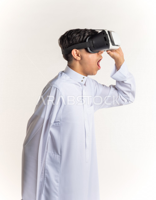 بورتريه شاب مراهق خليجي سعودي يقوم بالنظر من خلال نظارات الواقع الإفتراضي (في آر بوكس) بإيماءات وجه ويدين مختلفة , خلفية بيضاء
