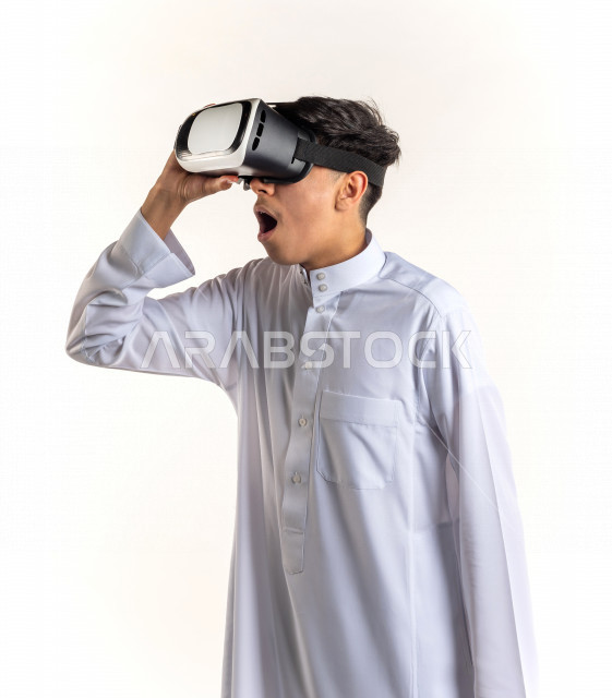 بورتريه شاب مراهق خليجي سعودي يقوم بالنظر من خلال نظارات الواقع الإفتراضي (في آر بوكس) بإيماءات وجه ويدين مختلفة , خلفية بيضاء