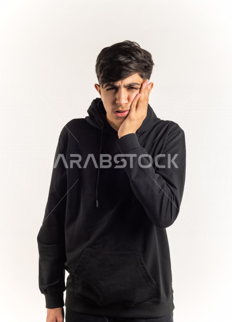 بورتريه شاب مراهق خليجي سعودي مريض بإيماءات وجه ويدين مختلفة تدل على الألم و الوجع في الأسنان ، خلفية بيضاء