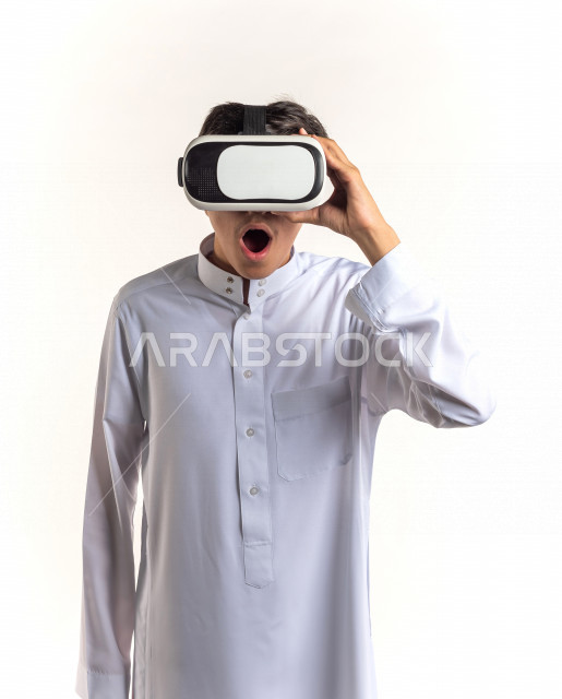 بورتريه شاب مراهق خليجي سعودي يقوم بالنظر من خلال نظارات الواقع الإفتراضي (في آر بوكس) بإيماءات وجه ويدين مختلفة , خلفية بيضاء
