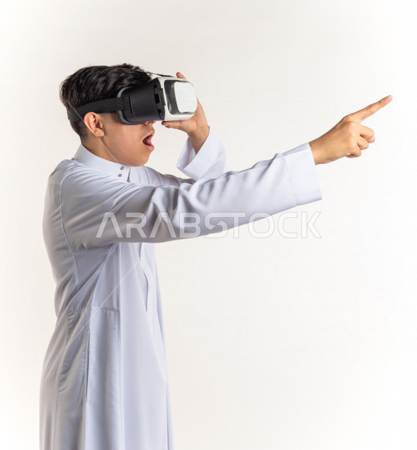 بورتريه شاب مراهق خليجي سعودي يقوم بالنظر من خلال نظارات الواقع الإفتراضي (في آر بوكس) بإيماءات وجه ويدين مختلفة , خلفية بيضاء