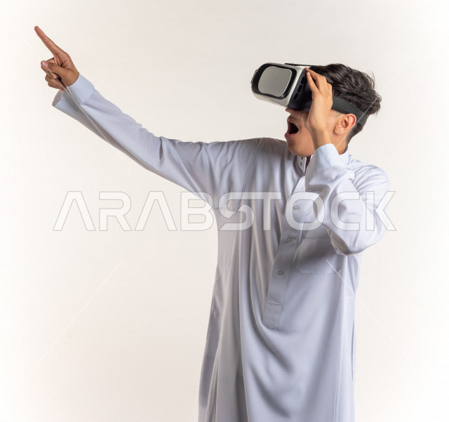 بورتريه شاب مراهق خليجي سعودي يقوم بالنظر من خلال نظارات الواقع الإفتراضي (في آر بوكس) بإيماءات وجه ويدين مختلفة , خلفية بيضاء
