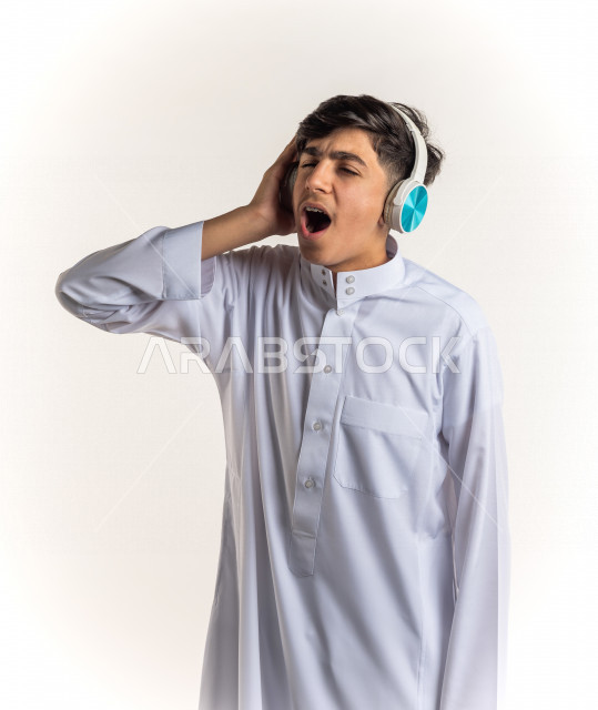 بورتريه شاب مراهق خليجي سعودي يضع سماعات على أذنيه بإيماءات وجه ويدين مختلفة تدل على الصوت العالي والتركيز , خلفية بيضاء