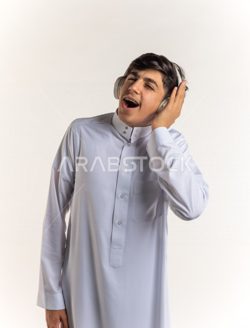 بورتريه شاب مراهق خليجي سعودي يضع سماعات على أذنيه بإيماءات وجه ويدين مختلفة تدل على الصوت العالي والتركيز , خلفية بيضاء