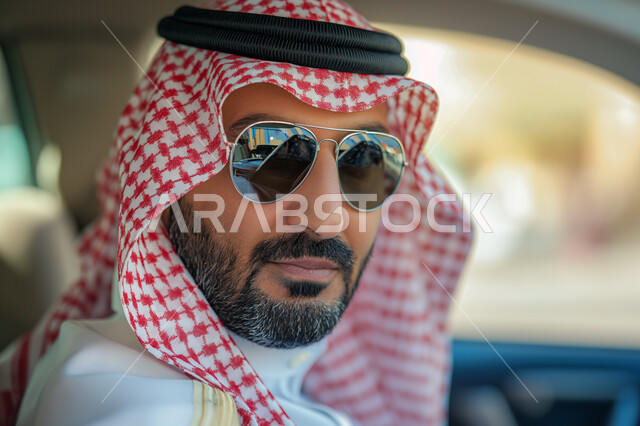 ايماءات الجدية والثقة بالنفس، المواصلات ووسائل التنقل في السعودية، مفهوم الأناقة والجولة، الاهتمام بالمظهر الخارجي، صورة مقربة لرجل عربي سعودي خليجي يرتدي الزي التقليدي والنظارة الشمسية يجلس بداخل سيارته