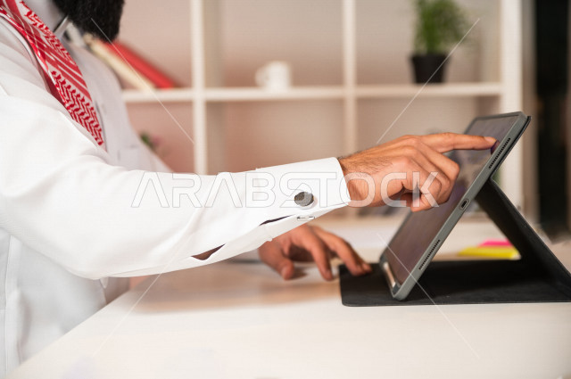 صورة مقربة لرجل اعمال سعودي خليجي يعمل في مقر الشركة ، يستخدم  جهاز تقني لانجاز مهامه  ،  يرتدي  لباس السعودي التقليدي  ، شركة سعودية ، عمل الخليج ، وظيفة مكتبية ، بيئة عمل
