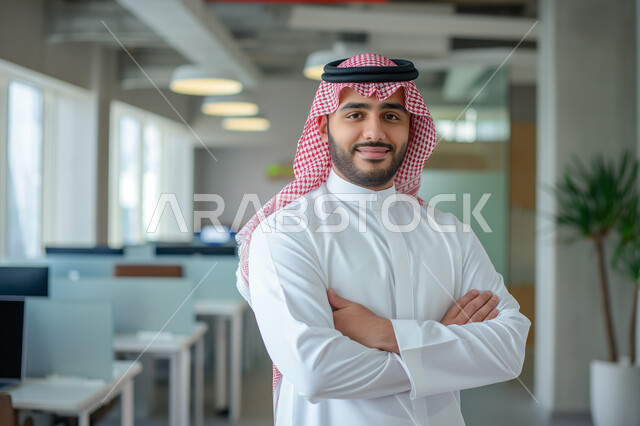 مفهوم الأناقة والرجولة والاهتمام بالمظهر الخارجي، شخصية سعودية مهمة يقف مكتوف اليدين، شاب عربي خليجي سعودي مبتسم يرتدي الزي التقليدي ينظر إلى الكاميرا بإيماءات الاعتزاز والثقه بالنفس, الوقوف في مكان العمل بسعادة
