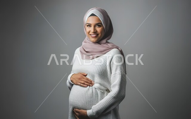 مفهوم الحمل والانجاب، بورتريه مقرب لإمرأة عربية خليجية سعودية محجبة تضع يدها على بطنها تنظر الى الكاميرا بإيماءات السرور، تعابير الحماس والشوق لانتظار المولود، الاهتمام بصحة الجنين، خلفية رمادية