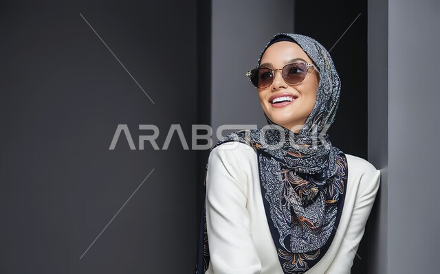 الاهتمام بالمظهر الخارجي، مفهوم الأناقة والشياكة و الأنوثة والنعومة، النظر إلى شيء ما بايماءات السعادة والسرور، أزياء محجبات عصرية حديثة، بورتريه مقرب لامرأة عربيه خليجيه سعودية محجبه ترتدي النظارة الشمسية, خلفية رمادية