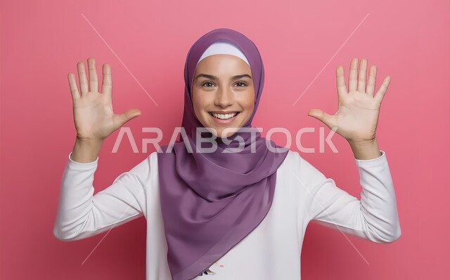 التعجب والاستغراب من شيء ما، بورتريه مقرب لفتاة مراهقة عربية خليجية سعودية مبتسمة تنظر الى الكاميرا وترفع يديها نحو الأعلى بإيماءات السرور، مفهوم النعومة والانوثة والاناقة، خلفية وردية