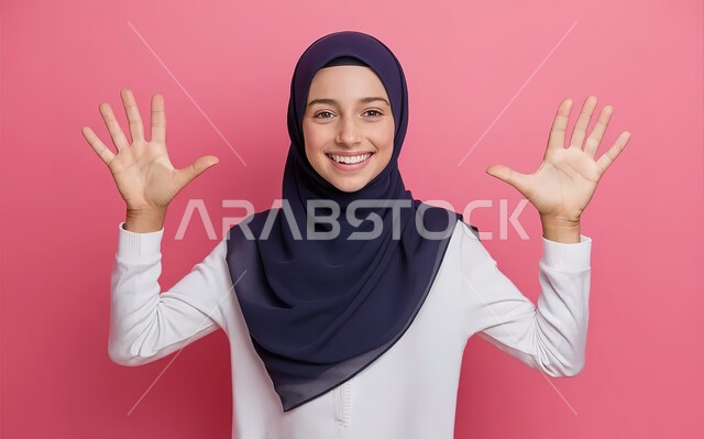 الاستغراب و التعجب من شيء ما، بورتريه مقرب لفتاة مراهقة عربية خليجية سعودية مبتسمة تنظر الى الكاميرا وترفع يديها نحو الأعلى بإيماءات السرور، مفهوم النعومة والانوثة والاناقة، خلفية وردية