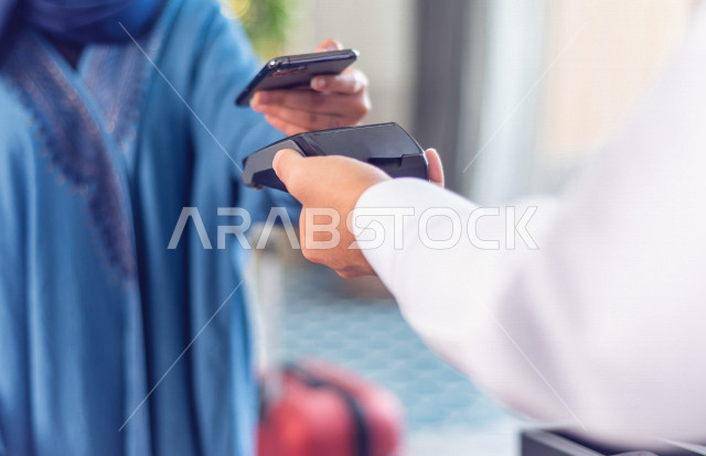 صورة مقربة لموظف إستقبال خليجي سعودي يعطي آلة الدفع لنزيلة سعودية محجبة في الفندق السياحي , تقوم بالدفع من خلال تطبيق بنكي عن طريق الهاتف المحمول, سياحة وفندقة, خدمة ومكتب إستقبال العملاء, إجراءات الحجز والإستفسار , سياحة وسفر