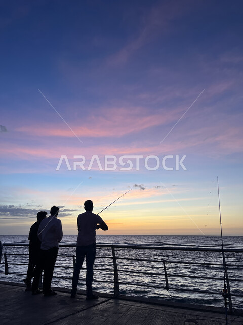 الكورنيش القديم المطل على البحر الأحمر، صورة ظلية لمجموعة من الشبان يتنزهون على ساحل مدينة جدة، أماكن سياحية في المملكة العربية السعودية، المناظر البحرية الهادئة، تأمل البحر وقت غروب الشمس، مفهوم صيد الأسماك، منظر الغيوم الوردية في السماء