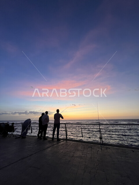 تأمل البحر وقت غروب الشمس، الكورنيش القديم المطل على البحر الأحمر، صورة ظلية لمجموعة من الشبان يتنزهون على ساحل مدينة جدة، أماكن سياحية في المملكة العربية السعودية، المناظر البحرية الهادئة، مفهوم صيد الأسماك، منظر الغيوم الوردية في السماء