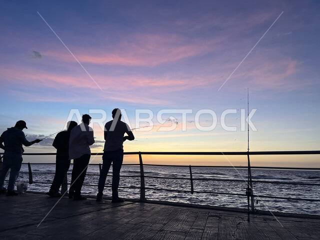 منظر الغيوم الوردية في السماء، الكورنيش القديم المطل على البحر الأحمر، صورة ظلية لمجموعة من الشبان يتنزهون على ساحل مدينة جدة، أماكن سياحية في المملكة العربية السعودية، المناظر البحرية الهادئة، تأمل البحر وقت غروب الشمس، مفهوم صيد الأسماك