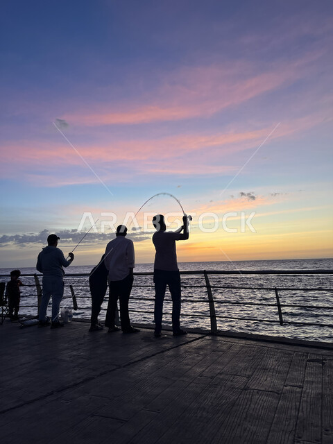 الكورنيش القديم المطل على البحر الأحمر، صورة ظلية لمجموعة من الشبان يتنزهون على ساحل مدينة جدة، أماكن سياحية في المملكة العربية السعودية، المناظر البحرية الهادئة، تأمل البحر وقت غروب الشمس، مفهوم صيد الأسماك، منظر الغيوم الوردية في السماء