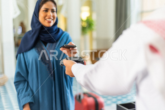 A close-up of a Saudi Gulf receptionist giving a payment machine to a veiled Saudi female guest in the tourist hotel, she pays through a bank application via a mobile phone, tourism and hotel, customer service and reception desk, reservation and inquiry p