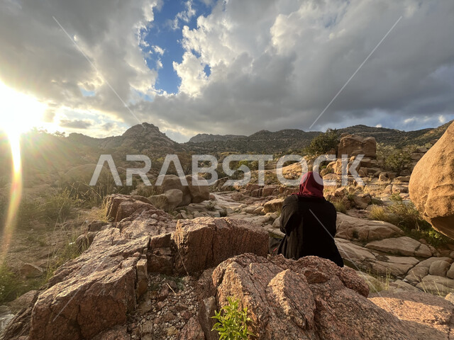 مفهوم تأمل المناظر الطبيعة، منظر الصخور والحجارة في الوديان، صورة من الخلف لشابة عربية خليجية سعودية تجلس في وادي الشفا في مدينة الطائف بالمملكة العربية السعودية، منظر الغيوم البيضاء في السماء، معالم أثرية سياحية، التضاريس والمرتفعات الجبلية