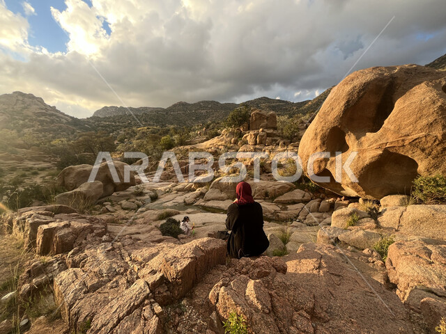 التضاريس والمرتفعات الجبلية، الاستمتاع بمشاهدة المناظر الطبيعية، منظر الصخور والحجارة في الوديان، صورة من الخلف لشابة عربية خليجية سعودية تجلس في وادي الشفا في مدينة الطائف بالمملكة العربية السعودية، منظر الغيوم البيضاء في السماء، معالم أثرية سياحية