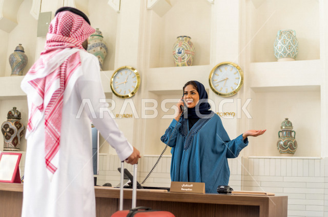 موظفة إستقبال خليجية سعودية محجبة مبتسمة ترحب بالنزيل السعودي وتتحدث على هاتف الفندق بإيماءات وجه ويدين مختلفة , سياحة وفندقة , خدمة ومكتب إستقبال العملاء , إجراءات الحجز والإستفسار , سياحة وسفر