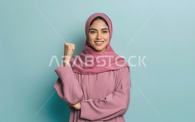 الانتصار و القوة  و التشجيع، إيماءات و تعابير السعادة و الفوز، بورتريه مقرب لإمرأة عربية سعودية خليجية محجبة مبتسمة ترتدي العباءة الوردية والحجاب ترفع يدها الى الاعلى وهي تقبض على قبضتها، 