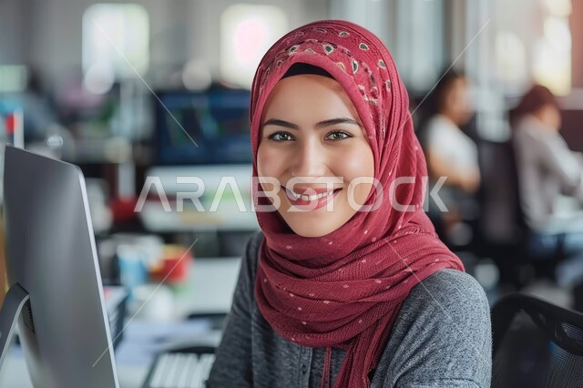 مهن ووظائف ادارية مكتبية، دمج العمل مع التقنية والتكنولوجيا، سيدة أعمال عربية خليجية سعودية محجبة تعمل على جهاز الحاسوب المحمول داخل المكتب تنظر الى الكاميرا بإيماءات السرور، إدارة شؤون الأعمال وتنظيمها عبر الاجهزة التقنية