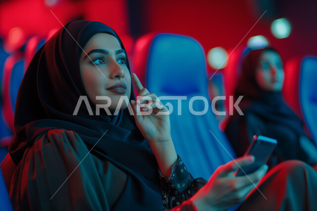 تجربه سينمائيه خياليه، صالات عرض رباعية الأبعاد, صورة مقربة لسيدة عربية سعودية خليجية محجبة ترتدي العباءة السوداء تجلس في صالة كي ماكس سينما تستمتع بالمشاهدة تمسك بيدها الهاتف المحمول، تعابير تدل على الفرح والسعاده، أنشطة ترفيهية متميزة