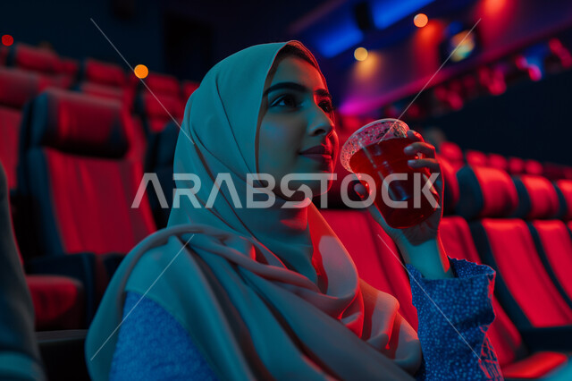 تجربه سينمائيه خياليه، صالات عرض رباعية الأبعاد, صورة مقربة لسيدة عربية سعودية خليجية محجبة ترتدي العباءة السوداء تجلس في صالة كي ماكس سينما تستمتع بالمشاهدة وشرب العصير، تعابير تدل على الفرح والسعاده، أنشطة ترفيهية متميزة