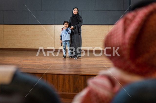 A saudi teacher's photo shoot honors a student in front of the audience at the school theater, a young Gulf child speaking with his microphone in his hand, an educational theater  