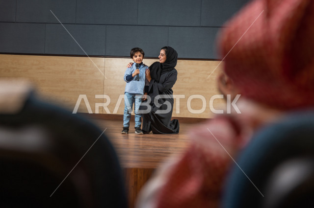 A saudi teacher's photo shoot honors a student in front of the audience at the school theater, a young Gulf child speaking with his microphone in his hand, an educational theater  