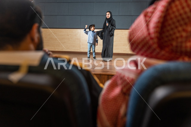 A saudi teacher's photo shoot honors a student in front of the audience at the school theater, a young Gulf child speaking with his microphone in his hand, an educational theater  