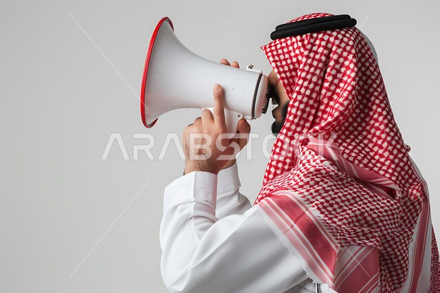Calling, announcing and drawing attention, using the microphone to announce something, a close-up portrait of a Saudi Gulf Arab man wearing the traditional dress and shemagh, holding a white loudspeaker in his hand, white background.