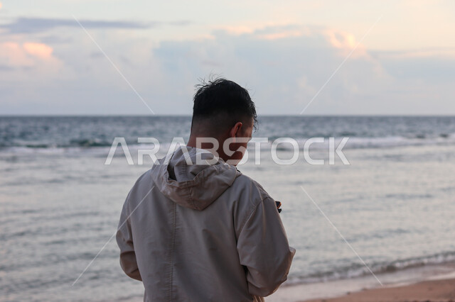  قضاء وقت ممتع بتأمل مناظر الطبيعة الهادئة، صورة من الخلف لشاب عربي خليجي سعودي يقف على الشاطئ الرملي، الاستمتاع بالنظر الى ساحل البحر، الواجهة البحرية والمناطق الطبيعية في المملكة العربية السعودية، النزهات والرحلات في فصل الصيف