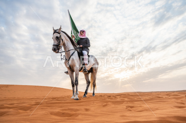 فارس سعودي خليجي يمتطي الخيل في صحراء السعودية  الخلابة ، يرفع بيده راية علم المملكة العربية السعودية  تحت سماء خلابة ملبدة بالغيوم ، الخيل العربي الأصيل، هواية ركوب الخيل ، الفروسية ، انشطة و فعاليات لركوب الخيل  ، اليوم الوطني السعودي