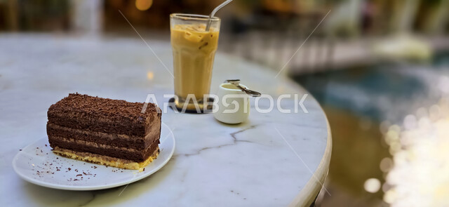 مشروب الايس لاتيه اللذيذ مع طبق من الحلويات الشهية على طاولة رخامية، الاستمتاع بالأجواء الهادئة في احدى كافيهات المملكة العربية السعودية، قطعة حلوى بالشيكولاته الشهية