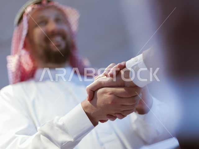 إلقاء التحية والسلام، صورة مقربة من الأسفل لأيدي رجلان عربيان خليجيان سعوديان يقومان بمصافحة بعضهما البعض، عقد صفقات جديده، التعاون بين الشركات العربيه في المملكه السعوديه