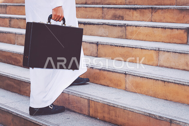 مفهوم الذهاب إلى العمل، صورة مقربة لموظف عربي خليجي إماراتي يقوم بصعود الدرج يمسك بيده حقيبة الملفات، مهن ووظائف مكتبية في الإمارات العربية المتحدة