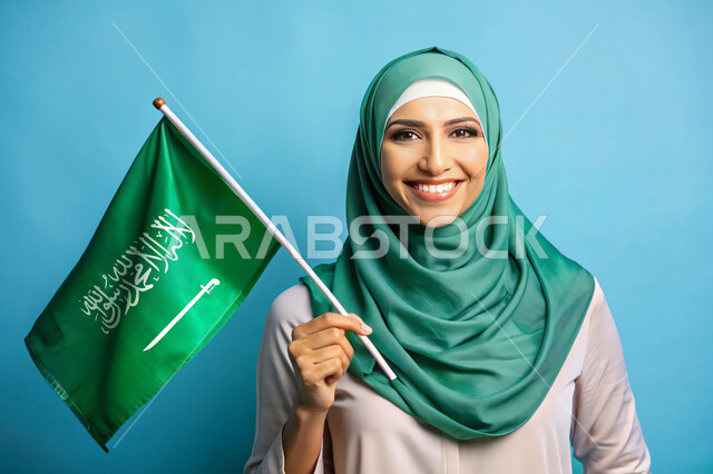 استذكار اليوم الوطني السعودي 23 سبتمبر، يوم العلم 11 مارس، الاحتفال بالأعياد والمناسبات الوطنية، رفع راية التوحيد والإسلام، بورتريه مقرب لامرأة عربية خليجية سعودية محجبة تحمل بيدها علم السعودية الاخضر، الفخر و الاعتزاز بالانتماء للوطن، خلفية زرقاء