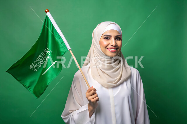 استذكار اليوم الوطني السعودي 23 سبتمبر، يوم العلم 11 مارس، الاحتفال بالأعياد والمناسبات الوطنية، رفع راية التوحيد والإسلام، بورتريه مقرب لامرأة عربية خليجية سعودية محجبة تحمل بيدها علم السعودية الاخضر، الفخر و الاعتزاز بالانتماء للوطن، خلفية خضراء، كروما
