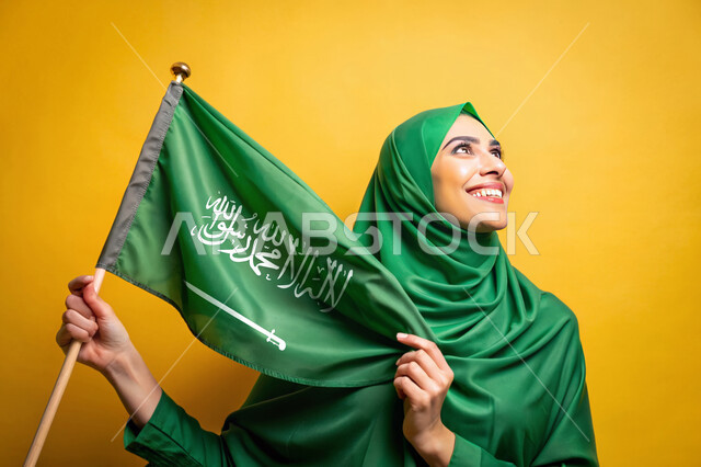 رفع راية التوحيد والإسلام، استذكار اليوم الوطني السعودي 23 سبتمبر، يوم العلم 11 مارس، الاحتفال بالأعياد والمناسبات الوطنية، بورتريه مقرب لامرأة عربية خليجية سعودية محجبة تنظر إلى الاعلى تحمل بيدها علم السعودية الاخضر، الفخر و الاعتزاز بالانتماء للوطن، خلفية صفراء